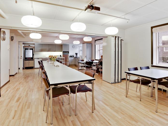 Lyst aflangt rum, som kan deles op med en foldedør. Rummet er indrettet med et langbord og mindre borde med stole omkring. Ved endevæggen et køkken med køleskab, ovn og opvaskemaskine.
