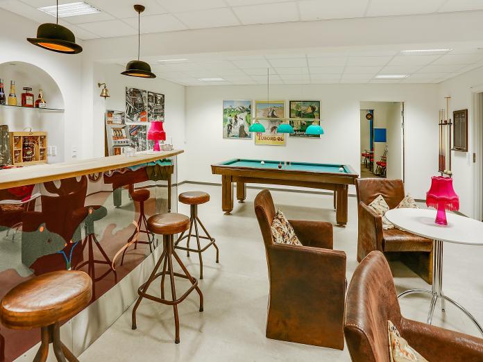 Fælles opholdsrum indrettet som bodega med billardbord, bardisk med barstole, spillemaskine og brune siddemøbler.