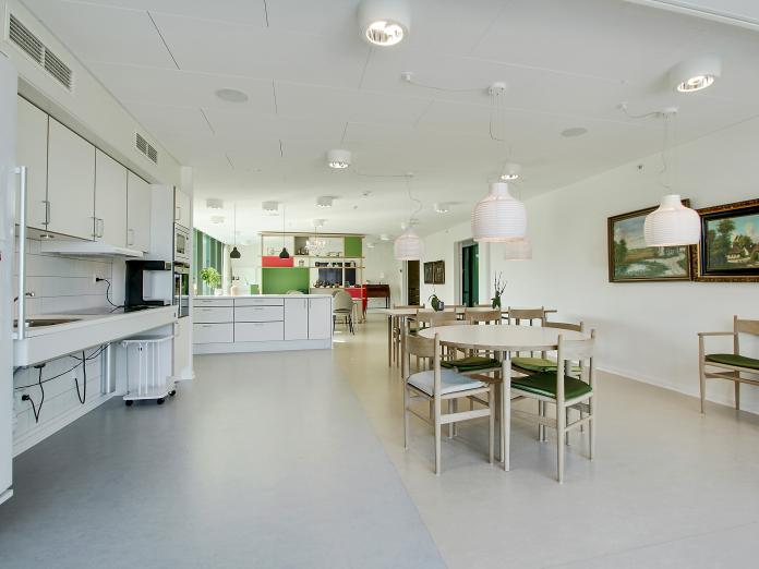 Stort, lyst fællesrum med køkken. Rummet er blandt andet indrettet med mindre borde med stole omkring og gamle malerier på væggene.