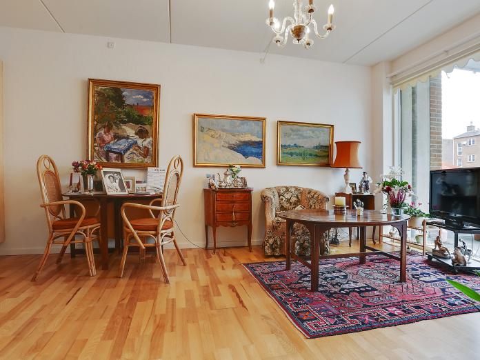 Stue med lyse vægge og lofter møbleret med personlige ejendele. Stuen har gulv til loft-vinduer med kig til privat altan.