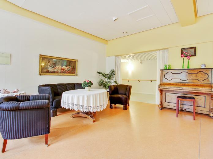 Hyggekrog med sofa og lænestole i mørkeblå brokadestof samt et mørkt sofabord med kniplingedug og potteplante. Op ad den ene væg står et klaver og på væggene hænger malerier.