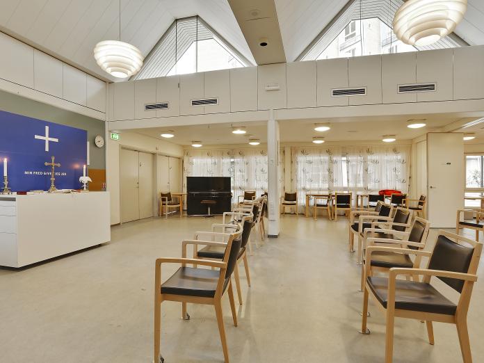 Stort, lyst fællesrum med højt til loftet. Rummet er indrettet med stole i rækker, et klaver og ad den ene væg er et alter med altertavle. 