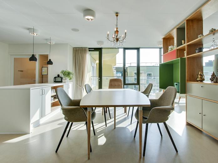 Fælles rum med gulv til loft-vinduer og dør ud til altan. Rummet er indrettet med aflangt spisebord med stole omkring og en stor reol. 