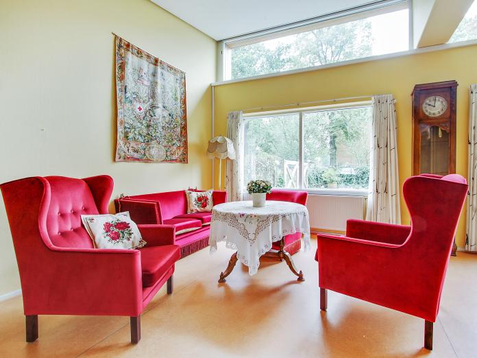 Hyggekrog med sofa og lænestole i rødt velour samt sofabord med kniplet dug og potteplante. Rummet er også indrettet med et bornholmerur, standerlampe med frynser og vægtæppe.