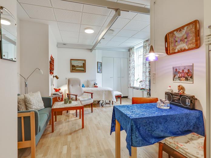 Lyst værelse set fra anden vinkel, hvorfra man kan se et lille spisebord med stole samt en sofa, sofabord, lænestol og bagerst i rummet en plejeseng og indbyggede skabe.