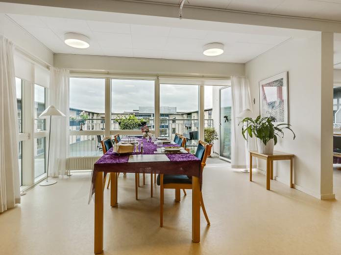 Hyggekrog med gulv til loft-vinduerne og udgang til stor altan. Krogen er indrettet med aflangt bord med stole, potteplanter, billede på væggen og standerlampe.