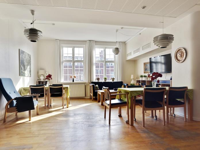 Lys fælles stue med sprossede vinduer. Stuen er indrettet med borde, stole, lænestol, sofa og sofabord samt væghængt fjernsyn.