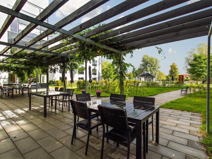 Pergola med klatregrønt over en fliseterrasse møbleret med sorte havemøbler og blomster i krukker.