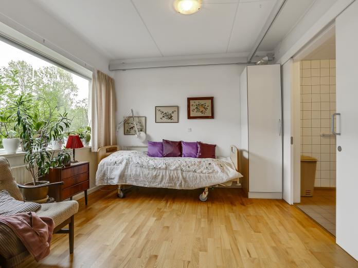 Soveværelse med seng, lænestol, billeder på væggene, vindue, et skab og en skydedør ud til badeværelset.