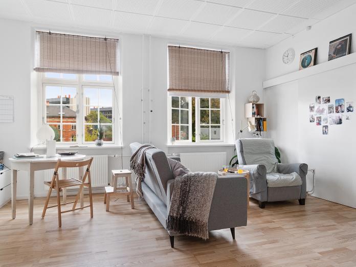 Lys stue med trægulve, højt til loftet og store, sprossede vinduer. Stuen er indrettet med personlige møbler som sofa, lænestol og sofabord samt spisebord med bænk og stole omkring. 