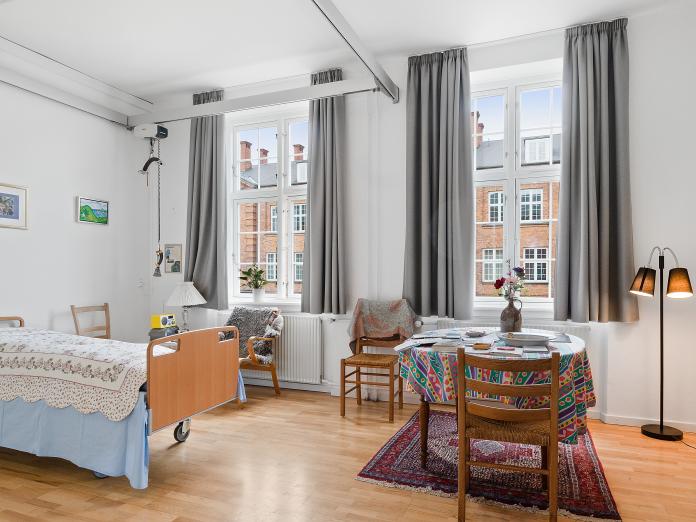 Værelse med store, sprossede vinduer. Rummet er indrettet med plejeseng og loftslift samt personlige møbler som bord, stole og standerlampe. På væggene hænger billeder, og der står blomster i vindueskarmen. 