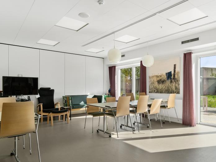 Fællesrum med store vinduespartier ud til en stor terrasse. Rummet er indrette med spiseborde og stole samt fjernsyn, lænestole og sofa.