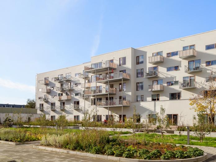 Moderne bygning med stålaltaner og vinduer i træ. Foran bygningen er en park med bænke, buske, bede og græs.