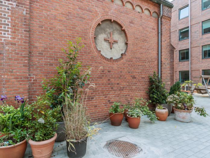 Hjørne af gårdmiljø, hvor der foran en rød murstensvæg er placeret mange krukker med blomster, græsser og grønne planter.