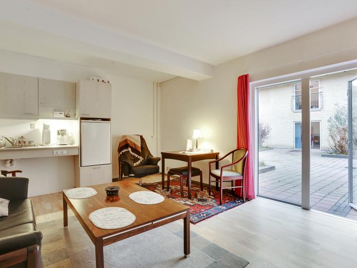 Stue med tekøkken med vask, kogeplader og køleskab samt skabe i lyst træ. Stuen har desuden gulv til loft-vindue og dør ud til fælles gårdhave. Stuen er møbleret med personlige møbler som sofa, lænestole og sofabord samt tæpper på gulvene.