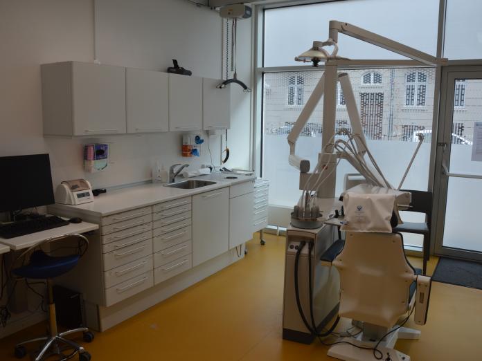 Tandklinik med tandlægestol, skabe, kontorstol og computer samt stort vindue.