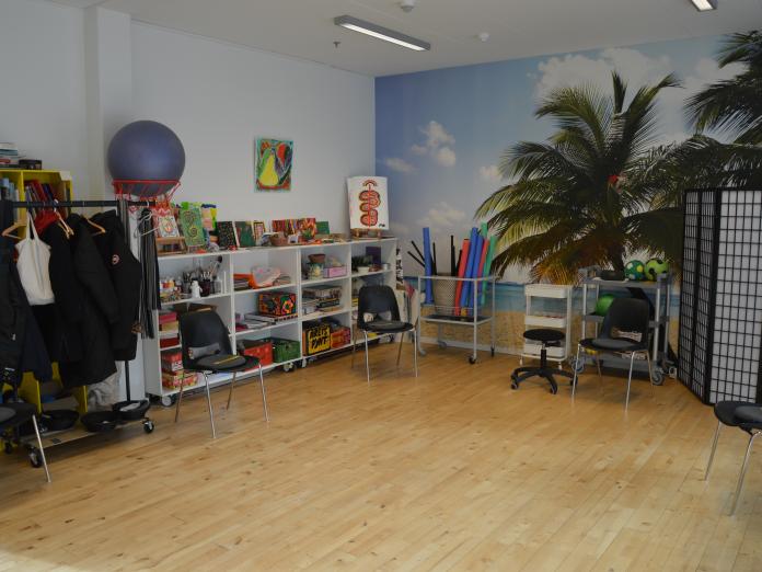 Kreativitetsrum med sorte stole og hvid reol med kreative værktøjer, blandt andet pensler og malerier. Rummet har et stort vægbillede med et motiv af en palmestrand.