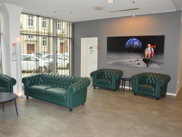 Lyst opholdsrum med gulv til loft-vinduer ud mod gaden. Rummet er indrettet med sofa, lænestole og siddebord samt et stort maleri på den ene væg.