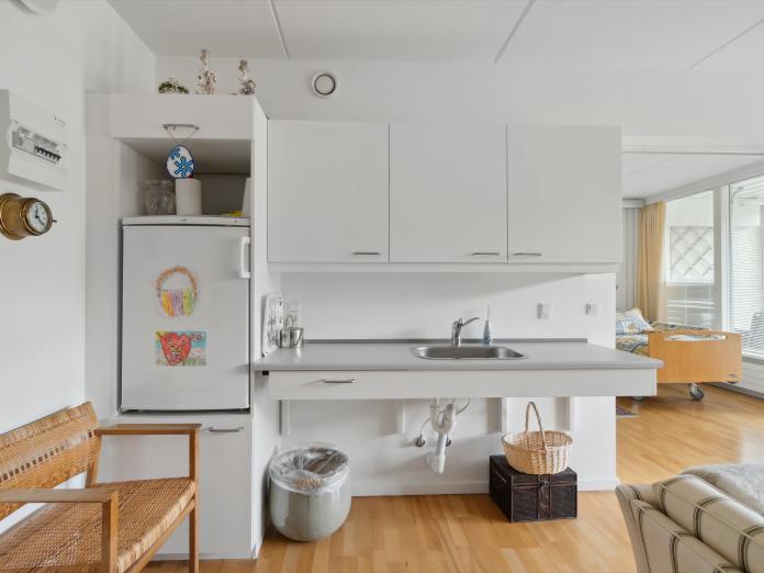 Tekøkken med køleskab med børnetegninger på, vask, bordplader, overskabe og ved siden af en lille bænk i flet.