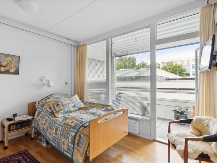 Plejeseng med sengebord, udgang til altan, stol og et lille tv, der hænger på væggen. 