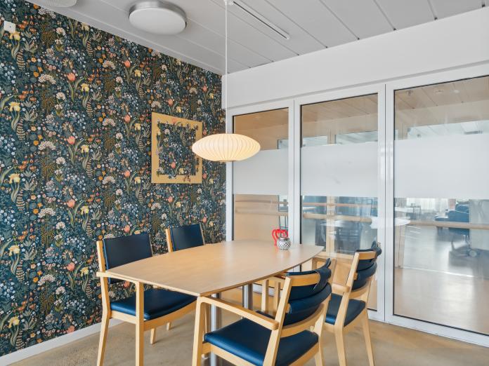 Fællesrum med blomstret tapet på væggen i mørke nuancer og et spisebord med fire stole.