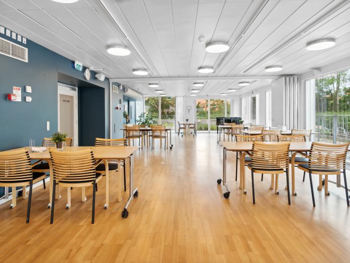 Fællesrum med mange spiseborde med stole, blåt på den ene væg og store vinduer.