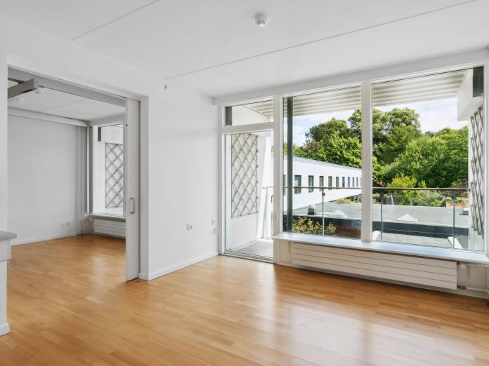 Umøbleret stue med kig til soveværelset samt altan, bygninger, træer og buske.