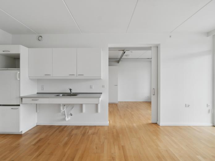 Umøbleret stue med tekøkken og kig til soveværelse gennem en åbne skydedør.