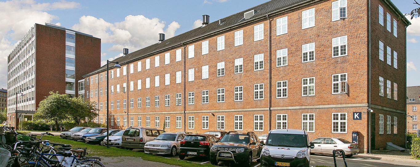 Ældre, rødt firetagers murstensbyggeri med sprossede, hvide vinduer. Foran bygningen er en vej, hvor der holder biler parkeret.