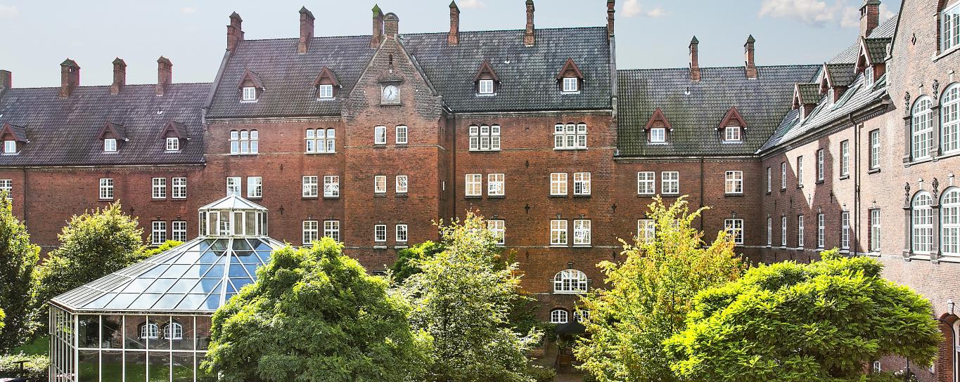 Ældre etagebyggeri i røde mursten og med hvide vinduer. Foran bygningen er et stort drivhus og mange store, grønne træer.