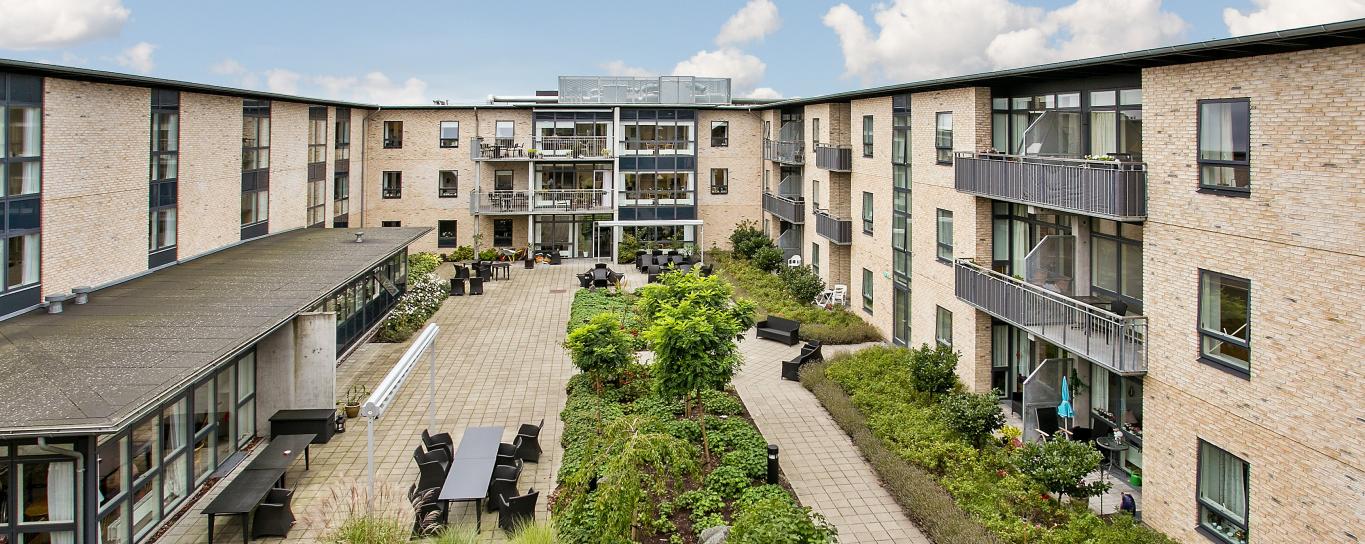 Lys treetagers bygning med altaner omkranser en indre gårdhave med bede, buske og træer.