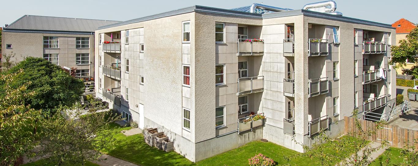 Moderne etagebyggeri med altaner. Foran bygningen er græsarealer med buske, blomster og træer.