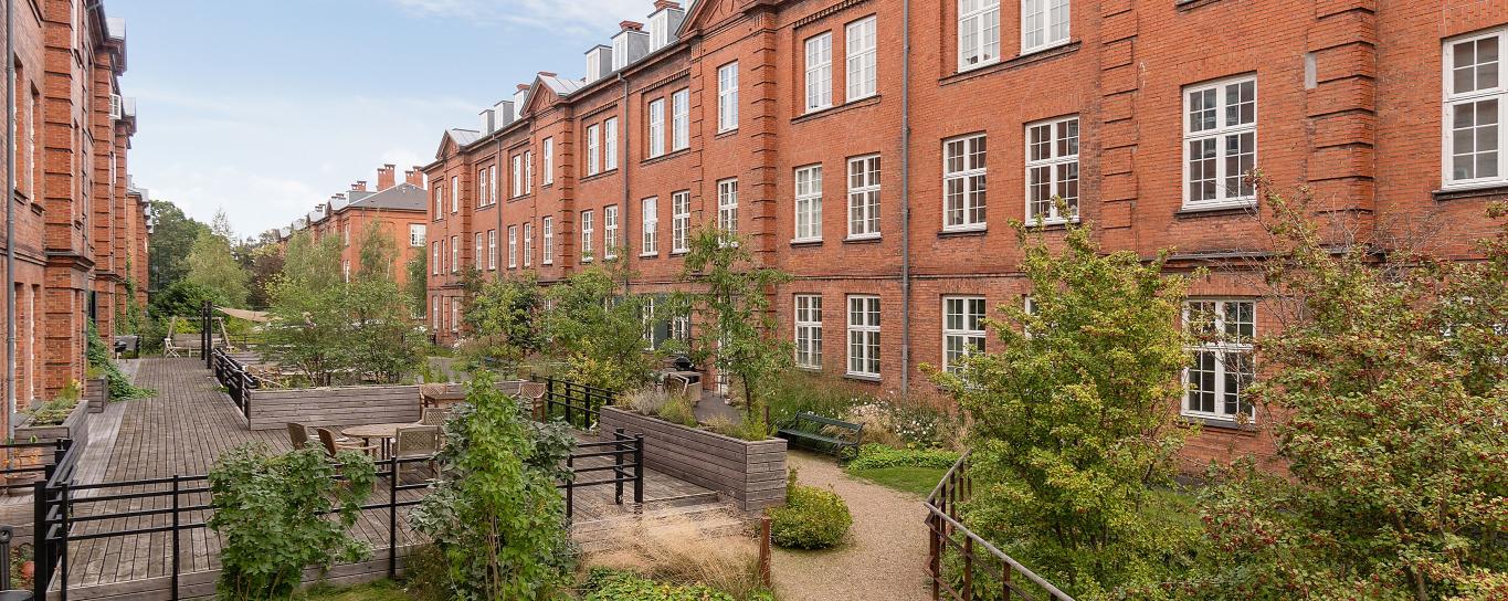 To ældre, røde murstensbygninger i fire etager. Mellem husene er en have med gangstier, buske, bede og træer samt bænke og havemøbler. 