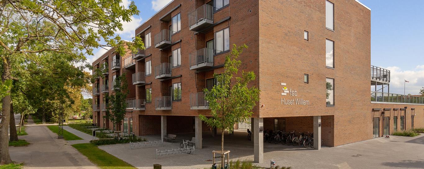 Moderne etagebygning i røde sten med stålaltaner og søljer i beton omkranset af fliser og grønne træer.