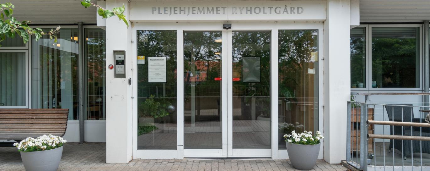 Indgangsparti til plejehjemmet Ryholtgård med skydelåger, krukker, bænke og grenene fra et træ. Over døren står nummeret 23.